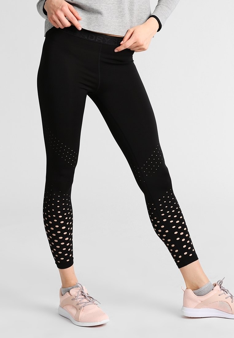 Osoba oblečená v černých legínách s perforovanými vzory a světle růžových teniskách se šedými ponožkami na jednobarevném pozadí.