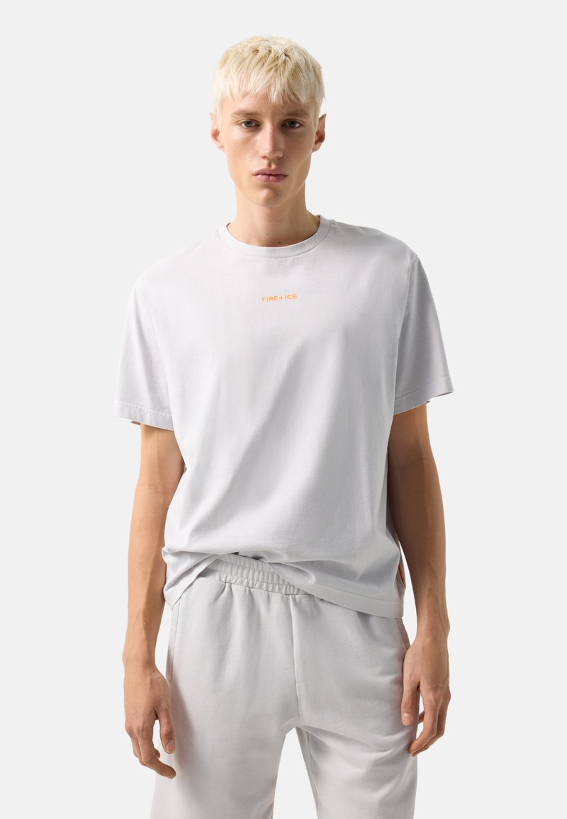 Homme blond portant un t-shirt blanc à manches courtes avec le texte orange « FIRE + ICE » et un pantalon blanc assorti, sur un fond clair uni.
