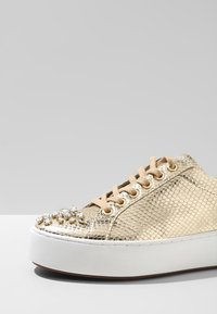 Zapatillas de piel de serpiente dorada con cordones beige, suela de plataforma blanca y un grupo de adornos de cristal transparente en la puntera.