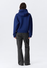 Μπλε-ναυτικό hoodie με μεγάλο κουκούλα και πέσμενους ώμους, σε συνδυασμό με γκρι τζιν φαρδιάς γραμμής, διαθέτει υφή υφάσματος και άνετη εφαρμογή.