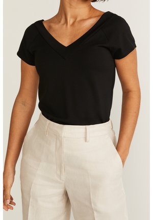 Woman wearing a black V-neck short-sleeve top tucked into high-waist beige trousers with one hand in pocket.