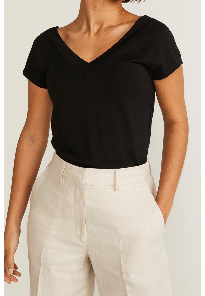 Femme portant un haut noir à col en V et manches courtes, rentré dans un pantalon beige taille haute, avec une main dans la poche.