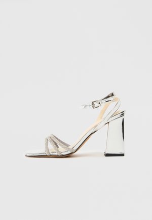 Sandale argentée à talon haut avec talon bloc, trois bandes en strass sur les orteils et une bride ajustable à la cheville avec boucle.