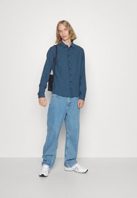 Camisa de cuadros azul y negra abotonada, jeans de mezclilla azul claro sueltos, zapatillas blancas y un pequeño bolso negro con correa para el hombro.