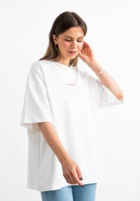 Witte oversized t-shirt met korte mouwen, voorzien van een klein rood logo. Soepele katoenstof met een relaxte pasvorm, gecombineerd met blauwe jeans.