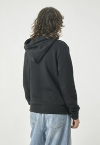 Schwarzer Kapuzenpullover mit einer großen, verstellbaren Kapuze. Hergestellt aus weichem Stoff, hat er gerippte Bündchen und einen geraden Saum. Getragen mit hellblauen Jeans.
