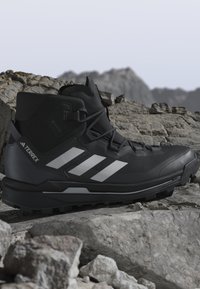 Sort Adidas Terrex vandrestøvle med hvide striber på klippefyldt bjergterræn, designet til udendørs og sporbrug.