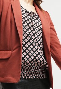 Blazer couleur rouille avec des épaules structurées, associé à un blouse noir et blanc à motif avec un ourlet froncé et un col rond.