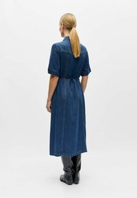 Femme avec une queue de cheval blonde portant une robe en denim bleu mi-longue avec une broderie florale sur les épaules et des bottes noires montant jusqu'aux genoux, debout.