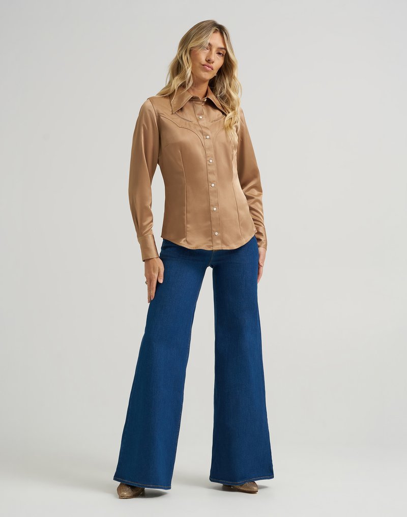 Camicia lunga in satin marrone con bottoni sul davanti e colletto a punta, abbinata a pantaloni in denim azzurri a gamba larga. Calzature lucide completano il look.