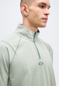 Νέος άνδρας που φοράει ανοιχτό πράσινο πουλόβερ με φερμουάρ έως τη μέση και ψηλό γιακά, με ένα μικρό οβάλ λογότυπο στο στήθος, κοιτάζοντας δεξιά.