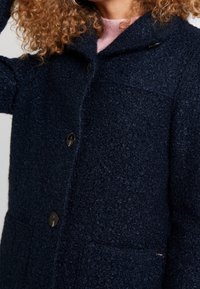 Cappotto in misto lana blu navy con finitura testurizzata, bottoni grandi, colletto sciallato e tasche laterali. Il design si concentra su calore e comfort.