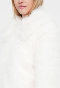 Cappotto in faux fur bianco con colletto alto, caratterizzato da una texture morbida e una finitura liscia. Il design mette in risalto calore e comfort.