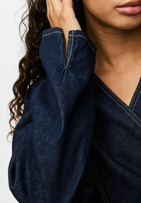 Tumšas džinsa topiņš ar V veida kakla izgriezumu, plašām piedurknēm un raksturīgām dzeltenām šuvēm. Gluda tekstūra ar viegli spīdīgu izskatu.