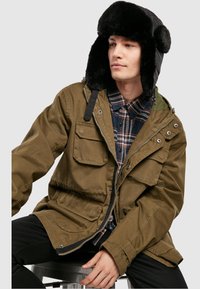 Olivově zelená parka s více předními kapsami, zipovým zapínáním a černou čepicí s umělou kožešinou. Model má pod ní na sobě kostkovanou košili.