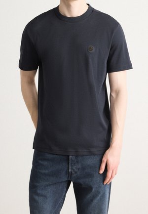 Mężczyzna noszący gładki czarny t-shirt z krótkim rękawem i okrągłym dekoltem, z małym ciemnym logo na lewej piersi oraz ciemnoniebieskie dżinsy.