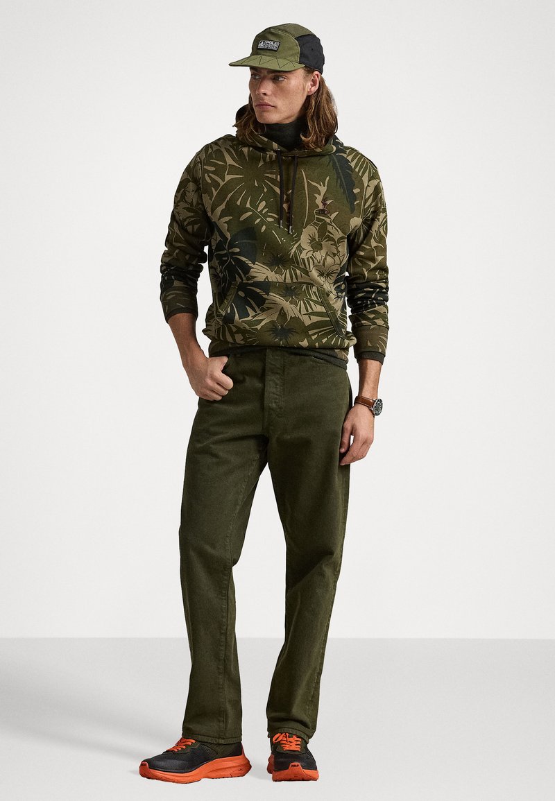 Kapuzensweatshirt mit grünem Blumenmuster, olivgrüne Hose und schwarz/orangefarbene Sneaker. Mütze in passendem Grün mit Logodetail.