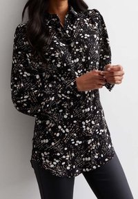 Mustapaita, jossa on valkoisia ja persikkaisia täpliä, napitettava kaulus ja pitkät hihat, joissa on hieman puffeja olkapäissä.