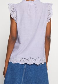 Vaalean violetti hihaton toppi, jossa on aaltoileva silmäreikäkuvio helman kohdalla ja röyhelöidyt hihansuut, valmistettu kevyestä, teksturoidusta kankaasta.