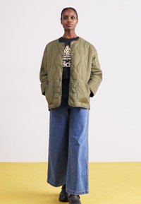 Olivno zeleni prešiti jopič s prsnim zapiranjem, nošen nad črno majico s tiskom in vparen z širokimi modrimi džins hlačami.