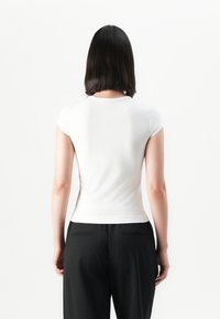 Femme aux cheveux raides et foncés portant un haut blanc ajusté à manches courtes et un pantalon noir, debout dos à l'appareil photo.