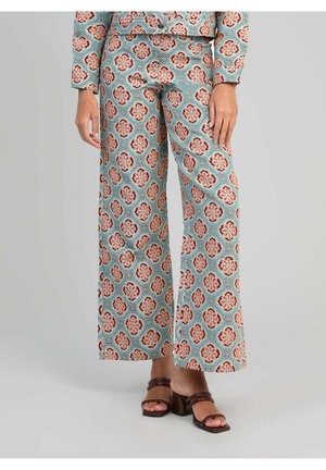 Femme portant un pantalon à jambes larges à motif floral bleu, rouge et beige, associé à des sandales à talons marron foncé.