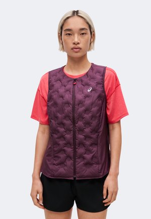 Young person with short hair wearing a purple quilted vest over a coral short-sleeve shirt and black shorts, standing against a white background.