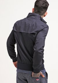 Svart bomberjacka med krage, dragkedja fram och ribbad fåll. Slät textur, figurnära design och ryggflik.