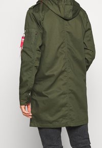 Parka verde larga con capucha, hecha de tela suave. Cuenta con bolsillos laterales, puños abotonados y un acento de etiqueta roja en la manga.