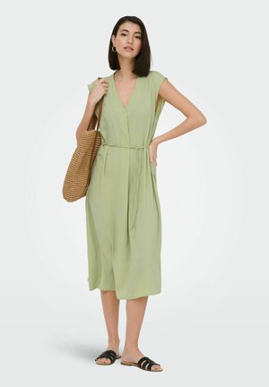 Žena stojaca v svetlo zelenej bezrukávovej midi šaty s opaskom v páse, drží pletenú kabelku cez rameno a má na nohách čierne sandále.
