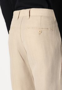 Pantalonii din in bej au un model în zig-zag, tăietură dreaptă și buzunare din spate cu închidere cu buton. Textură netedă și croi ajustat.