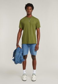 Maglietta verde a girocollo abbinata a pantaloncini di jeans. La modella tiene una giacca di jeans, indossa scarpe da ginnastica bianche e calzini a motivo.