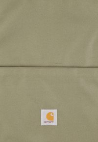 Gros plan sur un tissu khaki montrant une poche avec un patch logo Carhartt carré blanc arborant un emblème jaune et un texte rouge.