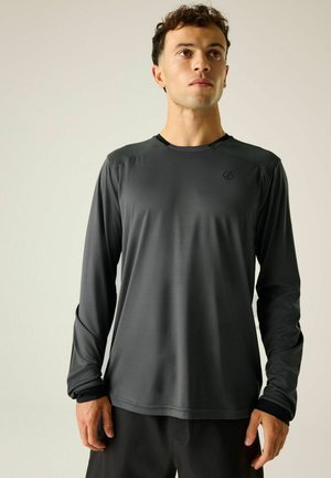 Junger Mann mit kurzen lockigen Haaren, der ein dunkelgraues langärmeliges Sportshirt trägt und vor einem hellen Hintergrund steht.