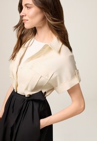 Camicia crema a maniche corte con tasche con bottone, abbinata a una gonna nera a vita alta con dettaglio fiocco legato in vita.