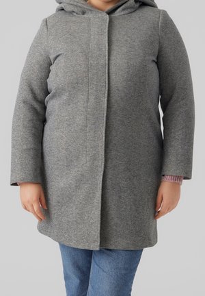 Enfant portant un manteau gris à capuche par-dessus un pull rose et un jean bleu, debout devant un fond clair uni.