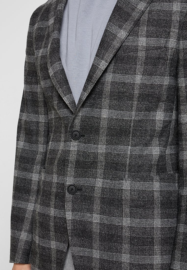 Grau und schwarz kariertes Blazer mit zwei schwarzen Knöpfen, getragen über einem hellgrauen Hemd.