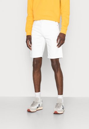 Mann trägt weiße kniehohe Shorts, gelbes langärmeliges Hemd, weiße Socken und graue Turnschuhe und steht vor einem schlichten Hintergrund.