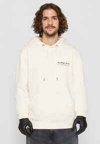 Krēmīga džemperis, kas izgatavots no mīkstas auduma, ar kanguru kabatu, regulējamu kapuci ar auklu un zaļu logotipa detaļu uz krūtīm.