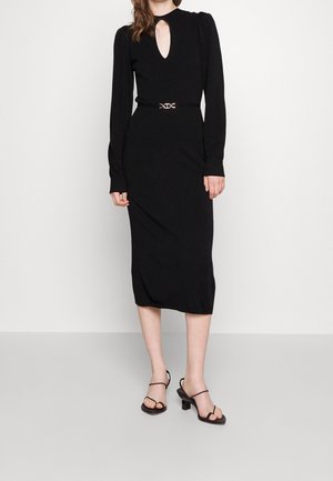 Woman wearing a black long-sleeve midi dress with a keyhole neckline and black heeled sandals, standing against a plain white background.