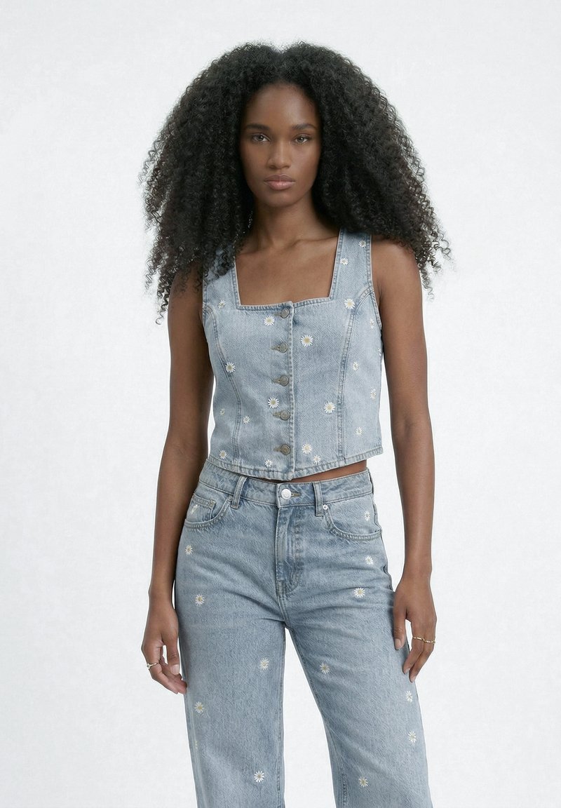 Femme aux longs cheveux bouclés portant un gilet en denim bleu clair et un jean, tous deux avec de petits imprimés marguerites blanches, debout devant un fond uni.