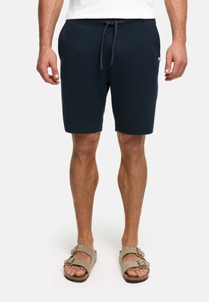 Mann trägt schwarze Shorts mit Kordelzug und beigen Sandalen mit Schnalle, steht vor einem schlichten hellen Hintergrund, Beine und unterer Torso sichtbar.