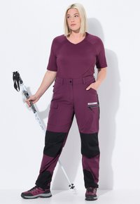 Venkovní turistický outfit, který zahrnuje fialové tričko s krátkým rukávem a kalhoty s černými akcenty, se zapínacími kapsami. Model drží trekingovou hůl a má na sobě turistické boty.