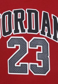 Le tissu rouge présente un grand texte noir "JORDAN" et des numéros gris "23", soulignés par une couture blanche, avec un motif texturé sur les numéros.