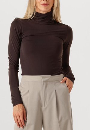 Brauner langer Pullover mit Rollkragen aus weichem Stoff, mit tailliertem Design, kombiniert mit leicht grauen, plissierten High-Waist-Hosen.