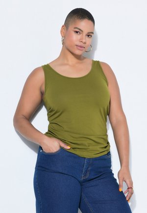 Donna con capelli corti che indossa orecchini a cerchio dorati, un top senza maniche verde oliva e jeans blu, in piedi con una mano in tasca.