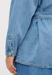 Light blue denim jacket featuring a dropped shoulder, cuff button details, and a belted waist with subtle texture in the fabric.