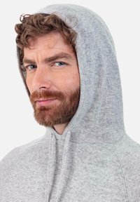 Sweat à capuche gris en maille avec capuche à cordon. Présente une texture douce et une coupe légèrement ample, mettant en valeur les coutures aux épaules.