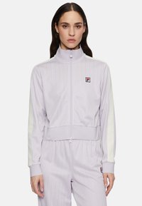Lila zip-up trainingsjack met verticale streepjes, witte schouderaccenten en een klein logo-opdruk op de borst. Zachte stoftextuur.