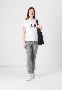 White t-shirt with Mickey and Minnie Mouse graphic, gray sweatpants, and white sneakers. Model holds a black tote bag.